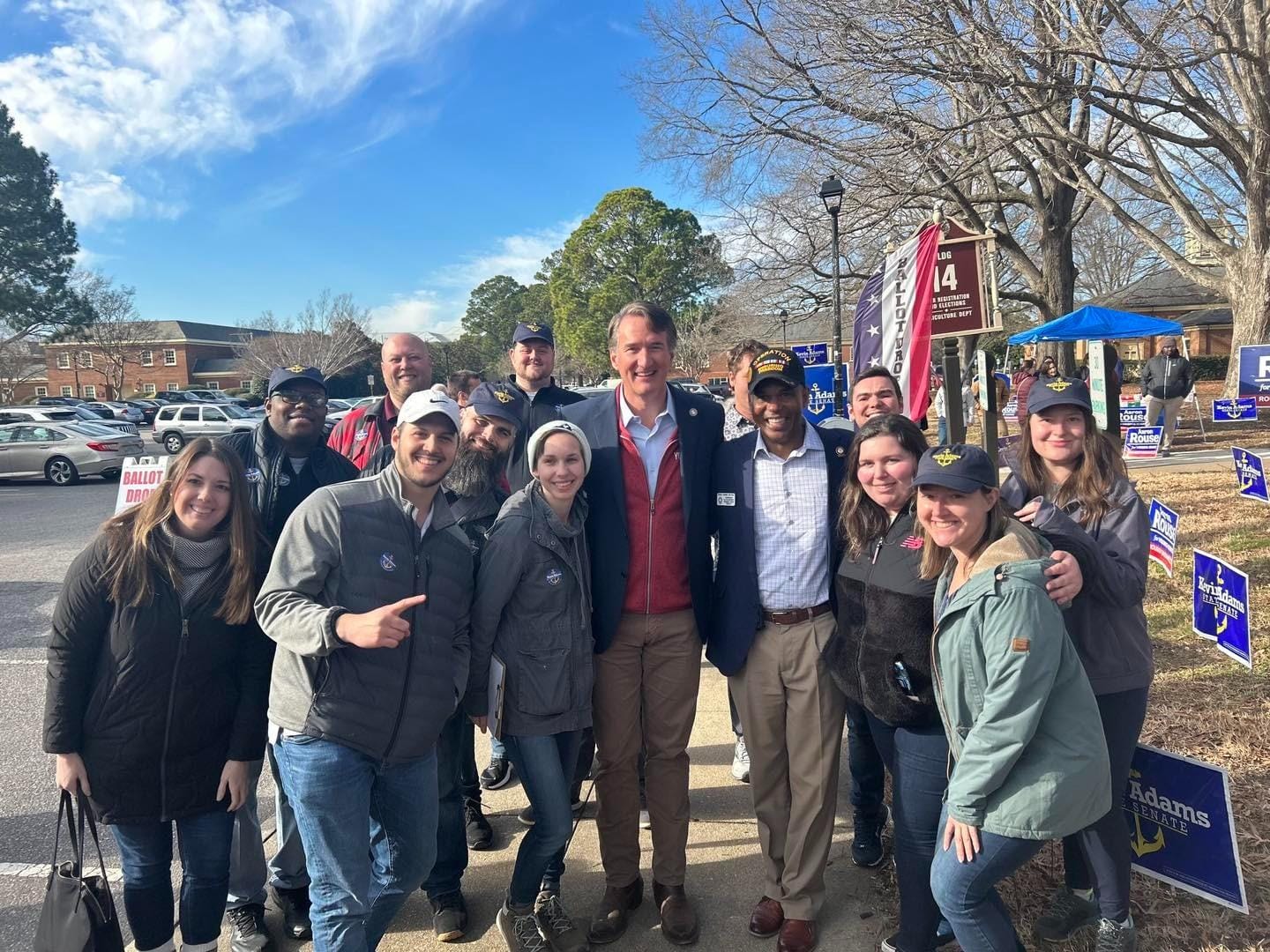 A group of people, consisting of men and women, in casual clothing gather around two central figures, likely for a photo o...