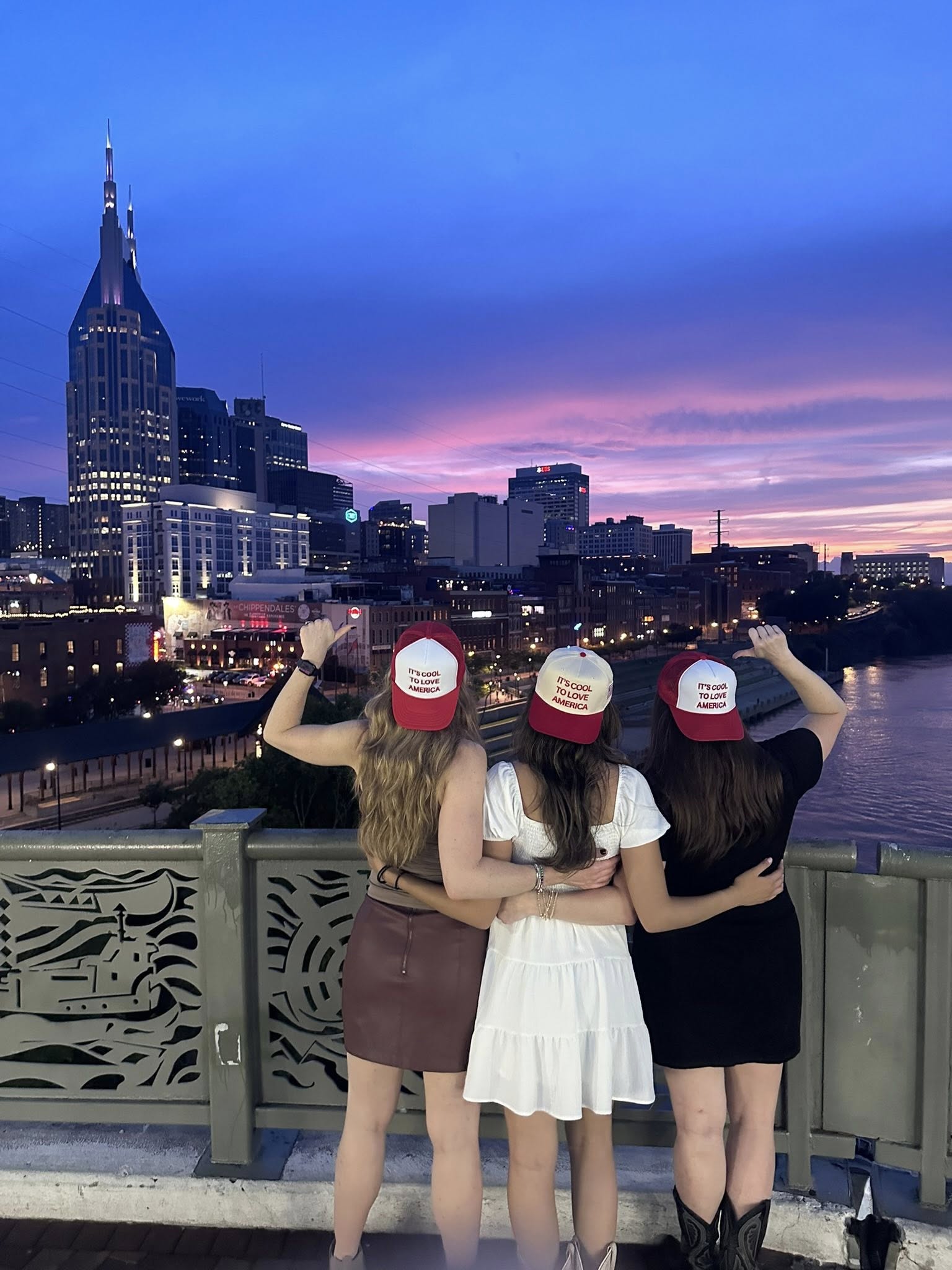 Three women, each wearing a hat that reads 'It's Cool to Love America,' standing on a bridge with Nashville, Tennessee's s...